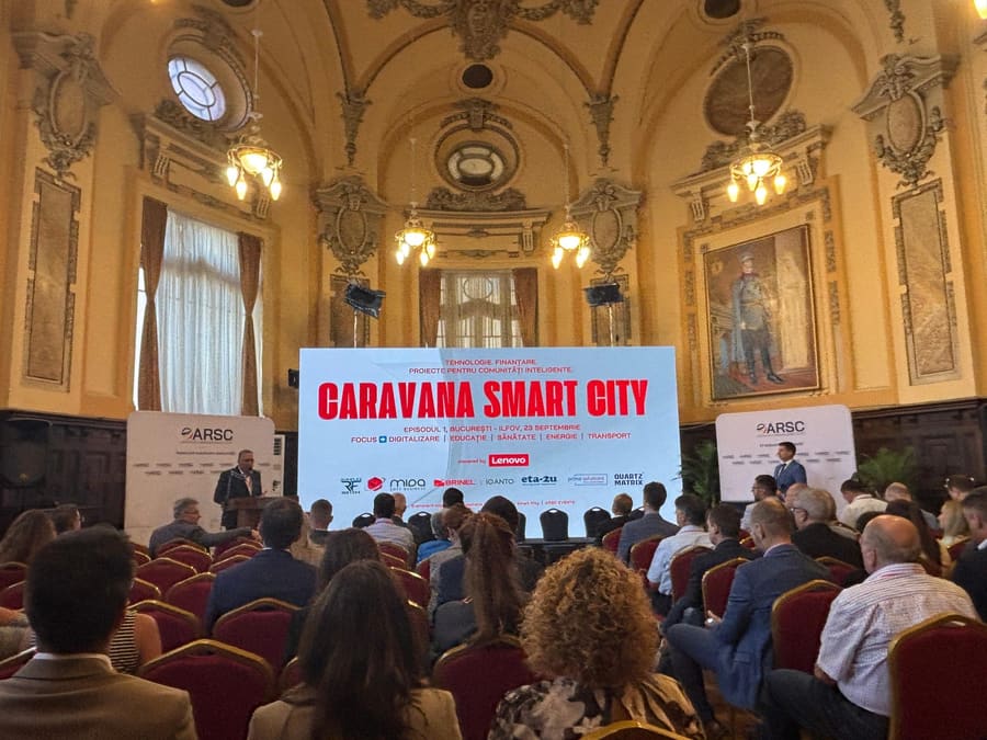 Sală plină cu public așezat, în fața unui ecran Caravana Smart City.