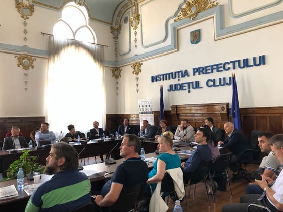 Sală oficială cu drapele și participanți la masă, la Instituția Prefectului Județul Cluj.
