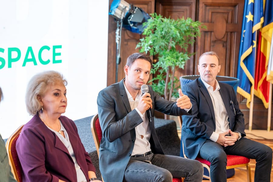 Trei vorbitori într-un panel, unul dintre ei răspunzând la microfon.
