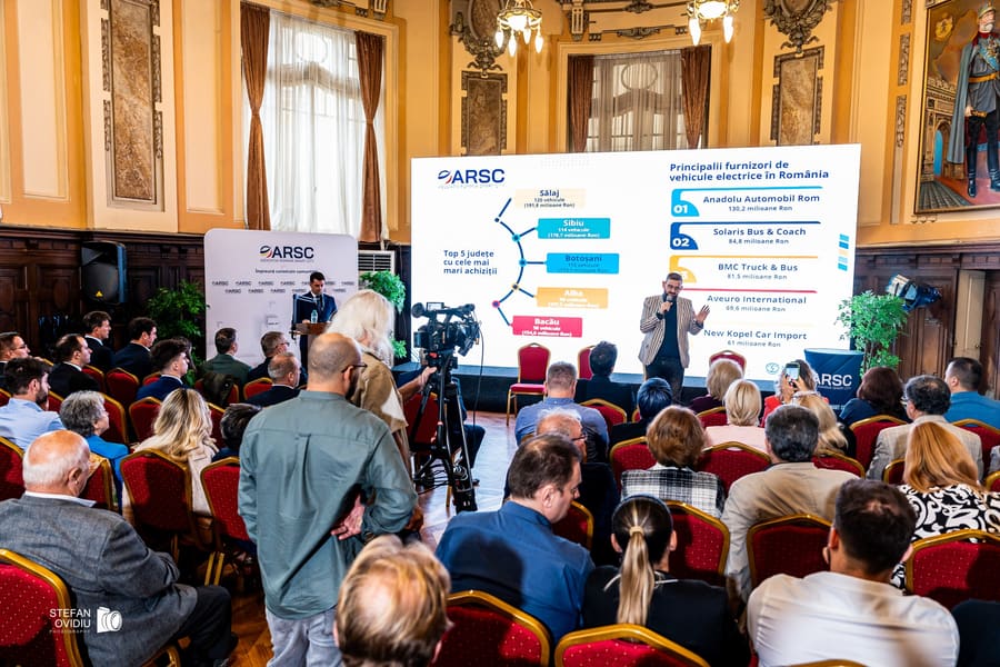 Public așezat pe rânduri urmărind o prezentare proiectată, cu o cameră de filmat în plan apropiat.