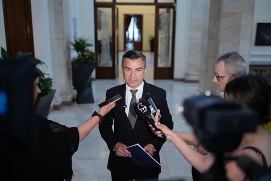 Vorbitor în fața microfoanelor de presă, cu cameră de filmat alături.