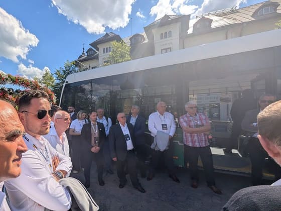Participanți adunați în jurul unui vehicul de transport public, în aer liber.