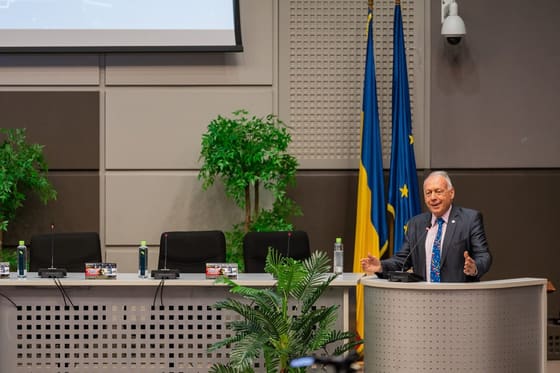 Vorbitor la pupitru, într-o sală oficială cu drapele.