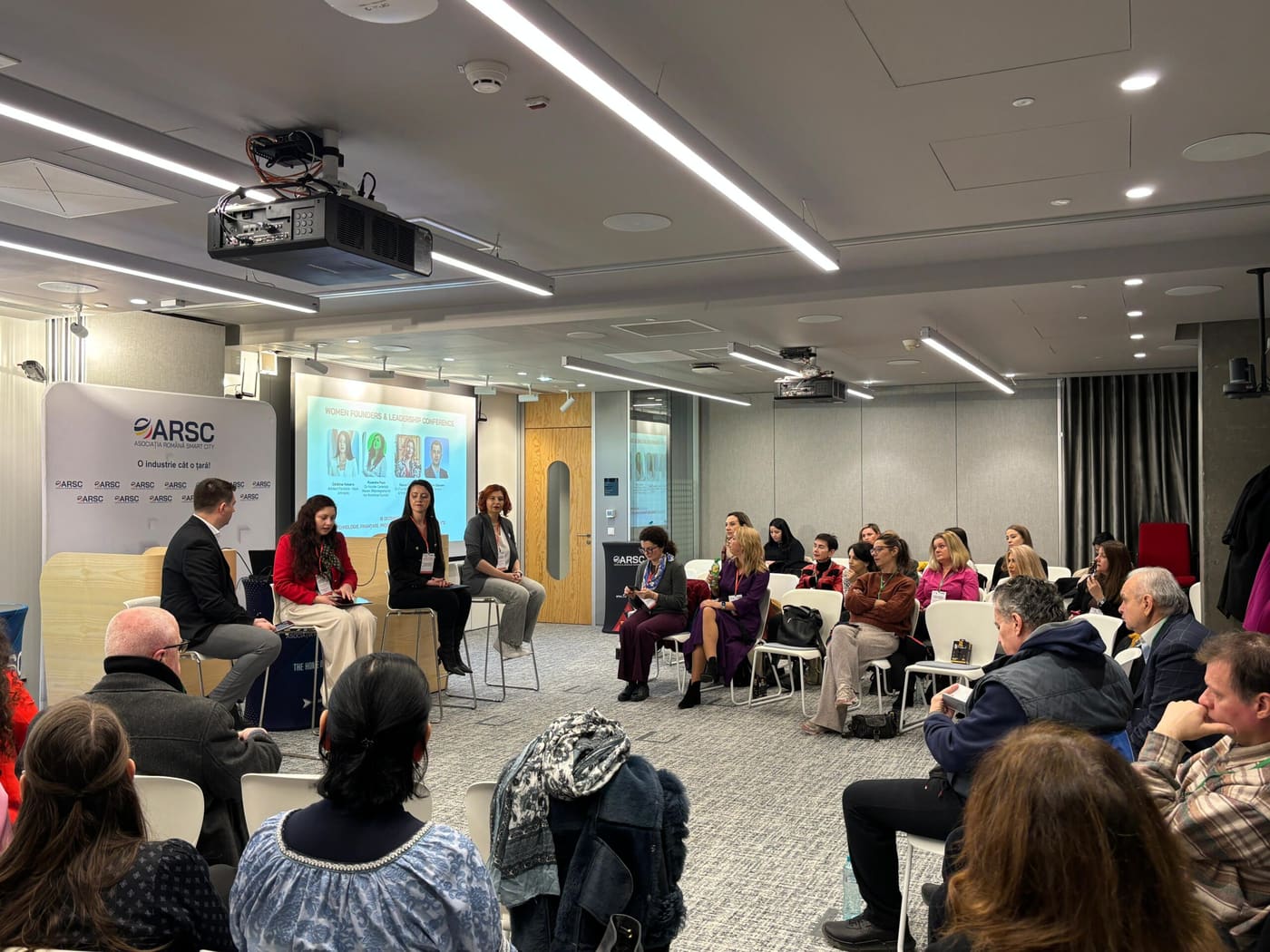 Sală de conferință cu un panel pe scaune înalte și participanți așezați în semicerc.